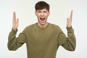 Young man, unhappy guy with brunette hair, piercing and bristle. Wearing khaki color sweater. Shouting angry with hands lifted. Watching at the camera isolated over white background photo