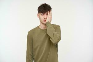 Young man, tired looking guy with brunette hair, piercing and bristle. Wearing khaki color sweater. Touching hes face with palm, exhausted. Watching at the camera isolated over white background photo