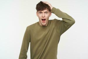 Portrait of confused, adult male with brunette hair, piercing and bristle. Wearing khaki color sweater. Touching hes had in shock of what he sees. Watching at the camera isolated over white background photo