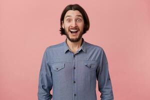 Photo of happy cheerful amazed bearded young man with long combed dark hair in denim shirt, heard the good news, was delighted, with open mouth, isolated over pink background.