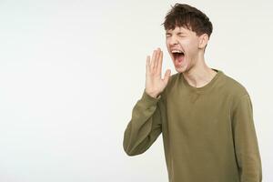 Young man, cool guy with brunette hair, piercing and bristle. Wearing khaki color sweater. Shout loudly with hand next to hes mouth. Stand isolated, copy space at the left, over white background photo