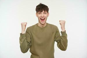 Teenage guy, happy looking man with brunette hair, piercing and bristle. Wearing khaki color sweater. Lifting up hes fists in excitement. Watching at the camera isolated over white background photo