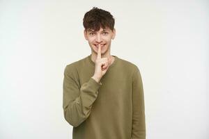 Young man, cool guy with brunette hair, piercing and bristle. Wearing khaki color sweater. Showing silence sign and smiling. Watching at the camera isolated over white background photo