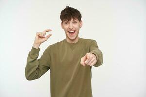 Cool looking male, handsome guy with brunette hair, piercing and bristle. Wearing khaki color sweater. Laughing, you were close. Pointing and watching at the camera isolated over white background photo