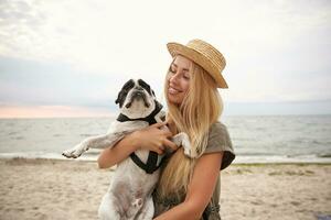 Positive young blonde woman with casual hairstyle posing over seaside on overcast, keeping her french bulldog and looking at him happily, wearing summer dress and boater hat photo