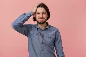 Portrait of young bearded man in denim shirt, hold his head, blowing cheeks and bites lips, forgot something important, made a mistake. Isolated over oink background. photo