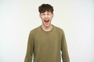 Cool looking male, handsome guy with brunette hair, piercing and bristle. Wearing khaki color sweater. Yelling hard with closed eyes, shout in anger. Stand isolated over white background photo