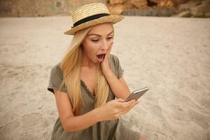 Horizontal shot of surprised pretty female with long blonde hair reading unexpected news on her mobile phone, looking at screen with wide mouth opened and keeping palm on her cheek photo