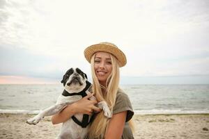 Photo of joyful pretty long haired female with blonde hair holding her funny dog on hands, looking in camera with cheerful smile while walking along beach on gray cloudy day