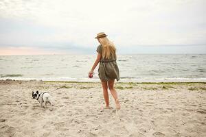 Outdoor photo of slim young female with long blonde hair wearing summer dress and boater hat, keeping her dog on leash while walking along beach on gray cloudy day