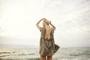 Bright outdoor photo of young slimwhite-headed female holding raised hands on her straw hat while enjoying the seaside view, wearing romantic green dress over beach background