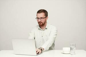 Concentrated young unshaved short-haired male in eyewear typing text with his laptop and looking attentively on screen, sitting over white background photo