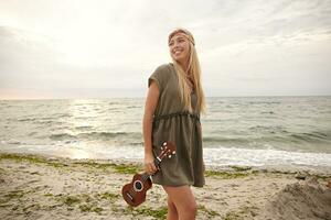 Outdoor photo of young beautiful white-headed female dressed in summer clothes holding ukulele and smiling widely while looking aside, isolated over seaside