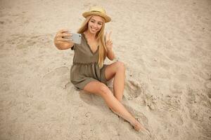Photo of charming blonde woman with long hair sitting over seaside in summer dress and straw hat, holding mobile phone and making selfie, raising hand with victory sign and smiling happily to camera
