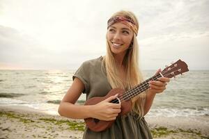 Outdoor photo of young charming white-headed woman in headband smiling happily while playing on small guitar, walking over seaside on bright warm day