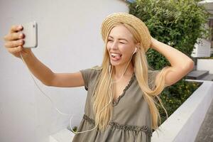 Outdoor shot of attractive young lady making selfie with her smarthone, winking and showing tongue, wearing casual linen dress and straw hat photo