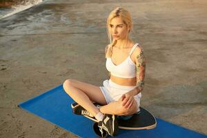 Young beautiful long haired blonde woman with tattooes sitting on wooden board over seaside promenade and looking on rising sun, wearing top and shorts photo