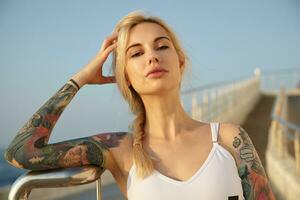 Close-up of beautiful tattooed young woman with long blonde hair combed in braid posing over seaside view, wearing white top, leaning head on raised hand and looking to camera with calm face photo