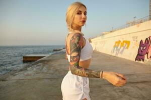 Portrait of attractive young tattooed female with long blonde hair combed in braid, posing over seaside promenade in sporty clothes, looking to camera with calm face and raising hands up photo