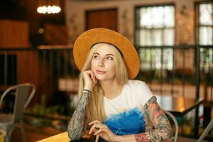 foto de bonito joven largo peludo mujer con casual peinado sentado a mesa en cafetería, mirando aparte afirmativamente mientras esperando para su orden, vistiendo blanco y azul camiseta y marrón sombrero