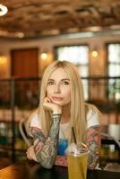 Portrait of beautiful blonde long haired female with tattooes leaning chin on raised hand and looking a camera with calm face, posing over restaurant interior in casual clothes photo