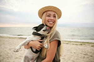 Joyful young pretty lady with blonde long hair posing over seaside on overcast with her french bulldog on hands, looking aside and smiling cheerfully, wearing casual clothes and straw hat photo