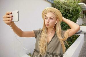 Attractive young woman with blond long hair in wicker hat walking along the street on sunny day, making faces and taking selfies on her phone photo