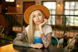 Indoor shot of puzzled blonde female with tattooes with posing over modern interior, sitting at table in cafe and leaning chin on raised hand, looking at camera with folded lips photo