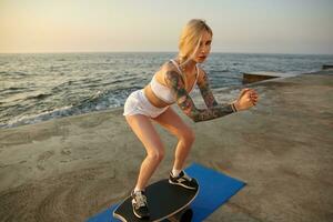 Photo of attractive slim young blonde woman with casual hairstyle standing over seaside view, wearing white top and shorts, helping to keep balance on board with raised hands