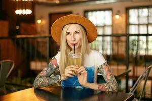 alegre rubia tatuado mujer con casual peinado Bebiendo limonada mientras esperando para amigos en ciudad cafetería, vistiendo blanco y azul camiseta y amplio marrón sombrero, mirando aparte con agradable sonrisa foto