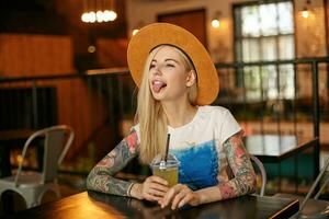 Funny shot of young blonde tattooed female with casual hairstyle sitting at table in restaurant, drinking juice while waiting for her order, looking aside joyfully and showing tongue photo
