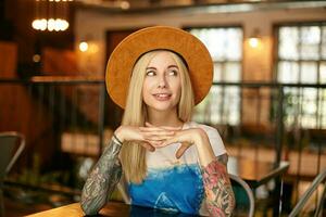 Portrait of pretty blonde female with tattooes looking aside with charming smile, raising folded hands while sitting over restaurant interior in casual clothes photo