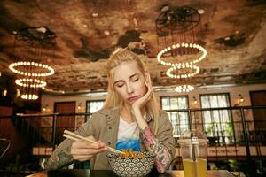 Horizontal shot of young attractive blonde woman with tattooes wearing beige shirt and white t-shirt, sitting at table over restaurant interior and having lunch, leaning chin on raised hand photo