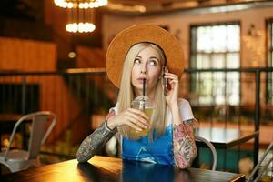 Horizontal shot of young blonde female with tattooes drinking fresh juice with straw, having lunch in city cafe, wearing white and blue t-shirt and brown hat photo