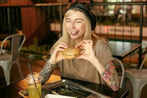 Indoor photo of joyful pretty woman with blonde long posing over cafe interior, having delicious lunch while sitting at table, keeping eyes closed with wide and cheerful smile