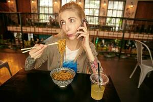 Indoor photo of pretty tattooed blonde woman with casual hairstyle eating asian food with wooden sticks, blowing on hot food while making call with her mobile phone