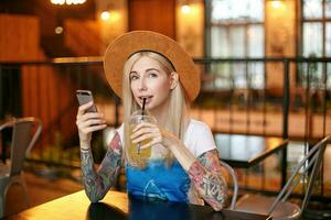 Positive tattooed blonde woman with casual hairstyle posing over modern indoor interior, sitting at table and drinking fresh juice, keeping mobile phone in hand and looking at camera wih light smile photo