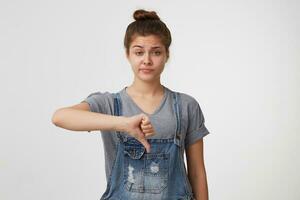 Unimpressed disliking attractive female student in denim overall, showing thumb down and smirking from displeasure, giving negative feedback about concert, frowning and standing over white wall photo