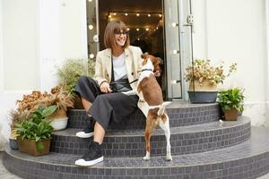 Street photo of attractive woman sitting on stairs, nearby is her lovely dog Jack Russell terrier, she looks happily, keeps dog's leash, petting her puppy, over street background