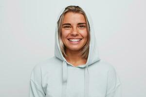 Portrait of cheerful pretty young woman with braces on teeth wears blue hoodie with hood feels happy and looks directly in camera isolated over white background photo