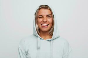 Closeup of happy playful young woman with braces on teeth wears blue hoodie with hood winks and shows tongue isolated over white background photo