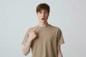 Portrait of sad amazed young man with short haircut in beige t shirt looks shocked and pointing at himself with finger isolated over white background photo