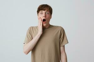 Closeup of astonished surprised young man in beige t shirt with short haircut and hands on cheek looks shocked and screaming isolated over white background photo