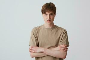 retrato de trastornado infeliz joven hombre con corto Corte de pelo en beige t camisa mira desesperado y en pie con brazos cruzado aislado terminado blanco antecedentes foto