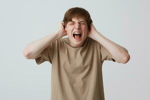 Closeup of crazy mad young man with closed eyes and braces on teeth in beige t shirt looks hysterical and screaming isolated over white background photo