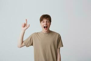 Portrait of happy excited young man with short haircut and braces on teeth in beige t shirt looks glad and pointing up isolated over white background photo