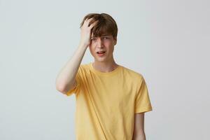 Portrait of desperate unhappy young man with hand on head and braces on teeth in yellow t shirt looks sad and having a headache isolated over white background photo