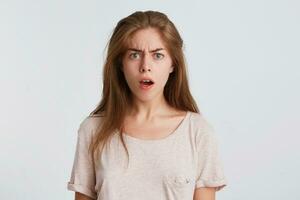 Closeup of angry shocked young woman with long hair wears t shirt standing with opened mouth and looks dazed isolated over white background photo