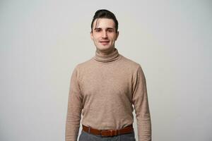Studio shot of young brown eyed stylish brunette man with trendy hairstyle posing over white background in beige roll-neck sweater and grey trousers, keeping hands along body photo