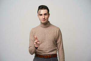 Handsome young brunette male hipster with stylish haircut raising hand and showing on camera with forefinger, keeping lips folded while posing over white background photo